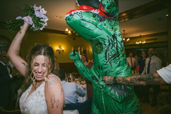 Una novia sonriente celebra junto a una persona disfrazada de dinosaurio.
