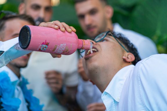 Joven bebiendo de una botella rosa mientras amigos lo observan en un ambiente festivo.